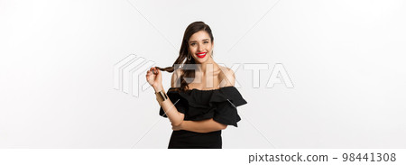 Beauty and fashion concept. Flirty woman in black dress, red lips, playing with hair and listening, looking at camera with interest, standing over white background 98441308