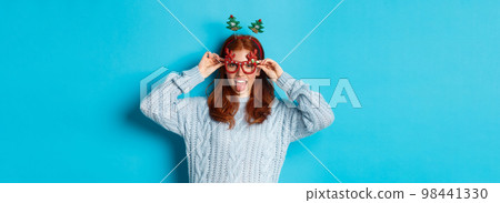 Winter holidays and Christmas sales concept. Beautiful redhead female model celebrating New Year, wearing funny party headband and glasses, smiling at camera 98441330
