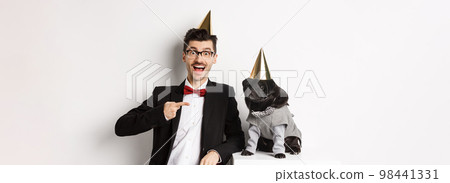 Cheerful man and cute black pug wearing party cones and suits, dog owner celebrating pet birthday, standing over white background 98441331