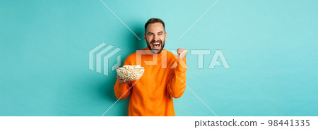 Cheerful handsome man in orange sweater, saying yes, cheering and rejoicing, eating popcorn and watching sports, making fist pump satisfied, standing over blue background Cheerful handsome man in orange sweater, saying yes, cheering and rejoicing, eating popcorn and watching sports, making fist pump satisfied, standing over blue background 98441335