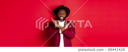 Angry Black man showing cross to stop something, looking furious and declining offer, standing in party clothes, red background 98441426
