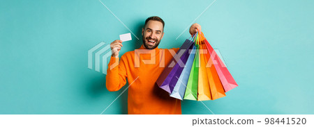 Happy aduly man showing credit card and shopping bags, standing against turquoise background Happy aduly man showing credit card and shopping bags, standing against turquoise background 98441520
