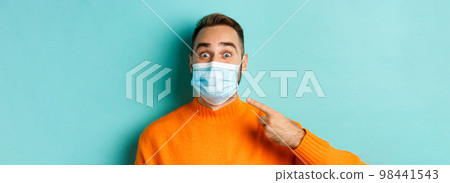 Covid-19, social distancing and quarantine concept. Close-up of cheerful man pointing finger at his medical mask, standing over turquoise background 98441543