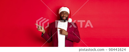 Christmas, party and holidays concept. Skeptical and unamused Black man looking with disdain, pointing fingers right at logo, standing in santa hat against red background 98441624