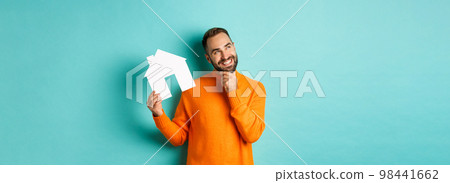 Real estate concept. Handsome adult man in orange sweater thinking about buying flat or renting, holding house maket and looking thoughtful, standing over blue background 98441662