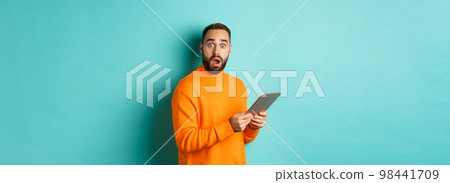 Image of male model in orange sweater using digital tablet, looking surprised, standing over light blue background Image of male model in orange sweater using digital tablet, looking surprised, standing over light blue background 98441709