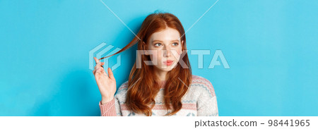 Close-up of thoughtful pretty redhead girl looking left, playing with hair strand and pondering, standing in winter sweater against blue background 98441965