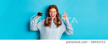 Happy redhead girl in sweater showing credit card and okay sign, recommending bank offer, standing over blue background Happy redhead girl in sweater showing credit card and okay sign, recommending bank offer, standing over blue background 98442552
