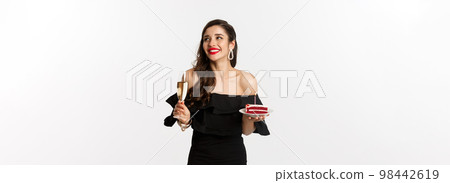 Celebration and party concept. Fashionable woman holding birthday cake with candle and drinking champagne, smiling and looking aside, standing over white background 98442619