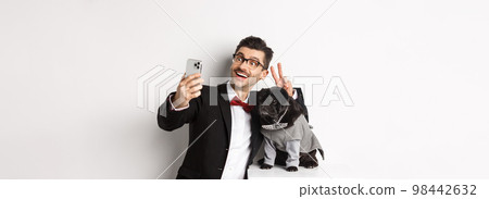 Cheerful dog owner in suit celebrating Christmas with dog, taking selfie on smartphone near cute black pug in costume, white background Cheerful dog owner in suit celebrating Christmas with dog, taking selfie on smartphone near cute black pug in costume, white background 98442632