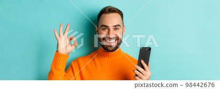 Close-up of bearded man holding smartphone and showing okay sign, recommending mobile app, standing satisfied over turquoise background Close-up of bearded man holding smartphone and showing okay sign, recommending mobile app, standing satisfied over turquoise background 98442676