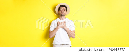 Concept of tourism and summer. Romantic man in straw hat looking nostalgic, close eyes and holding hands on heart, standing against yellow background Concept of tourism and summer. Romantic man in straw hat looking nostalgic, close eyes and holding hands on heart, standing against yellow background 98442781