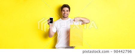 Concept of mobile banking and cashback. Young happy guy holding shopping bag and showing smartphone screen, yellow background 98442796