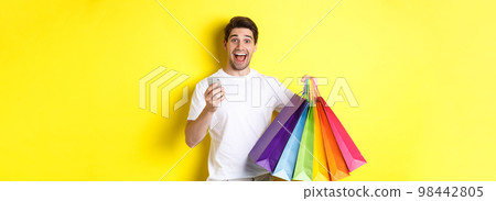 Image of happy man receive cashback for purchase, holding smartphone and shopping bags, smiling excited, standing over yellow background 98442805