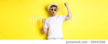 Concept of tourism and lifestyle. Happy guy tourist enjoying trip, rooting for you, fist pump and triumphing, going on journey in summer hat and sunglasses, yellow background 98442806
