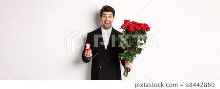 Handsome smiling man in black suit, holding roses and engagement ring, making a proposal to marry him, standing against white background Handsome smiling man in black suit, holding roses and engagement ring, making a proposal to marry him, standing against white background 98442860
