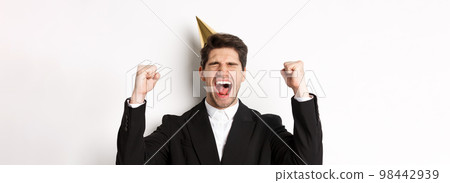 Close-up of happy good-looking man, wearing party hat and suit, raising hands up and rejoicing, celebrating new year, standing against white background 98442939