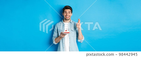 Hopeful man holding birthday cake and cross fingers, making wish, standing over blue background 98442945
