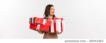 Holidays, celebration concept. Beautiful caucasian woman in elegant dress holding Christmas presents and smiling happy, standing over white background 98442963