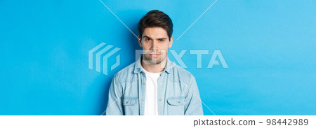 Close-up of suspicious and serious guy raising one eyebrow, looking at camera, standing against blue background Close-up of suspicious and serious guy raising one eyebrow, looking at camera, standing against blue background 98442989