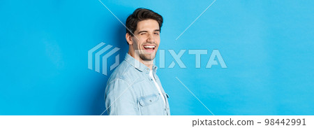 Happy and confident man turn head at camera, winking and smiling, standing over blue background Happy and confident man turn head at camera, winking and smiling, standing over blue background 98442991