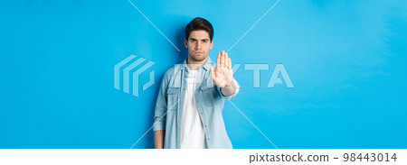 Serious man frowning and saying no, extending hand to shop stop sign, prohibit action, standing against blue background 98443014