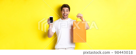 Surprised man holding shopping bag and showing smartphone screen, concept of mobile banking and app achievements, yellow background 98443018