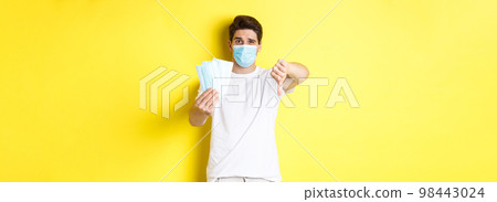 Concept of covid-19, quarantine and preventive measures. Man looking disappointed and showing thumb down, do not recommend bad medical masks, standing over yellow background 98443024