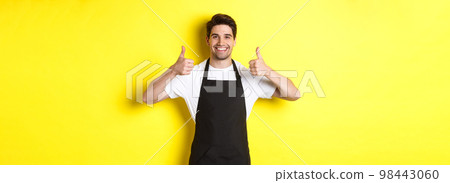 Smiling seller in black apron showing thumbs up, approve or like something, recommending cafe or store, yellow background 98443060