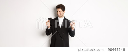Portrait of confident handsome businessman shopping online, holding credit card and smartphone, standing over white background 98443246