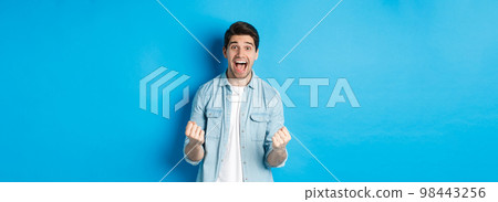 Excited handsome man winning, feeling lucky and rejoicing, celebrating victory and shouting yes, standing against blue background Excited handsome man winning, feeling lucky and rejoicing, celebrating victory and shouting yes, standing against blue background 98443256