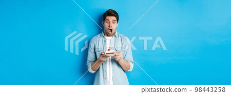 Man looking shocked at birthday cake, standing against blue background Man looking shocked at birthday cake, standing against blue background 98443258