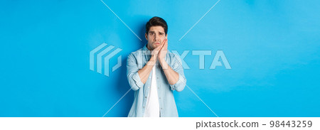 Portrait of man with toothache grimacing from pain and touching cheek, standing against blue background Portrait of man with toothache grimacing from pain and touching cheek, standing against blue background 98443259