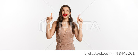 New Year, christmas and celebration concept. Happy elegant woman in party dress, laughing and looking up, pointing at top promo offer, white background New Year, christmas and celebration concept. Happy elegant woman in party dress, laughing and looking up, pointing at top promo offer, white background 98443261