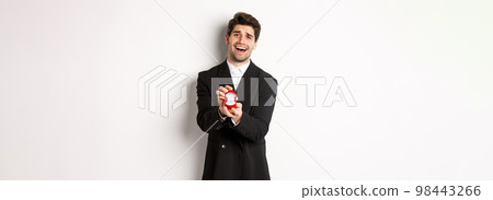 Portrait of handsome man in black suit, open box with wedding ring, making a proposal, asking to marry him, standing against white background 98443266