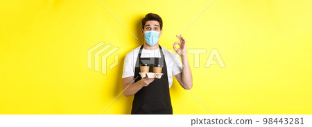 Concept of covid-19, cafe and social distancing. Barista in medical mask and black apron guarantee safety, holding takeaway cups of coffee and showing OK sign, yellow background Concept of covid-19, cafe and social distancing. Barista in medical mask and black apron guarantee safety, holding takeaway cups of coffee and showing OK sign, yellow background 98443281