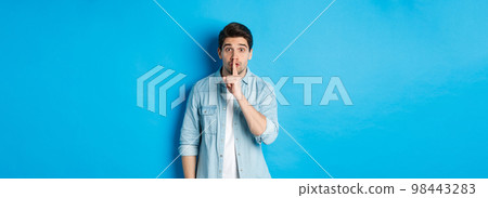 Portrait of excited man asking to keep quiet, showing hush taboo sign and looking nervously at camera, standing against blue background 98443283