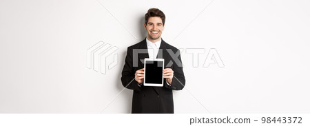 Portrait of handsome businessman in trendy suit, showing digital tablet screen and smiling, standing against white background 98443372