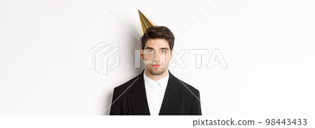 Close-up of handsome man in party hat and trendy suit, celebrating new year, standing against white background 98443433