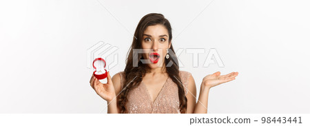 Close-up of attractive brunette woman with red lips, wearing dress, receiving engagement ring and looking surprise, standing over white background 98443441