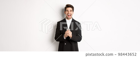 Portrait of happy good-looking man, wearing black suit, laughing from happiness and using mobile phone, standing over white background 98443652