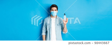 Concept of covid-19, pandemic and social distancing. Young man in medical mask showing hand sanitizer, standing against blue background Concept of covid-19, pandemic and social distancing. Young man in medical mask showing hand sanitizer, standing against blue background 98443667