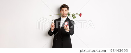 Romantic young man in suit making a proposal, holding rose in teeth and glass of champagne, showing engagement ring, asking to marry him, standing against white background 98443693