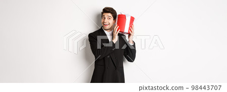 Concept of christmas holidays, celebration and lifestyle. Handsome man in black suit, shaking a box with gift, wondering whats inside, standing against white background Concept of christmas holidays, celebration and lifestyle. Handsome man in black suit, shaking a box with gift, wondering whats inside, standing against white background 98443707