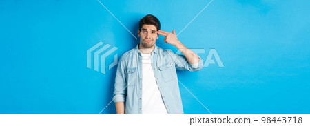 Tired and annoyed adult man making finger gun gesture near head, shooting himself from boredom, standing against blue background Tired and annoyed adult man making finger gun gesture near head, shooting himself from boredom, standing against blue background 98443718