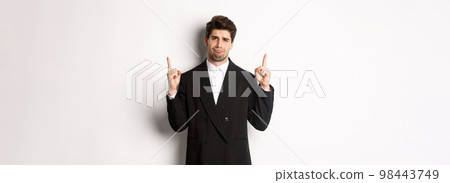 Portrait of reluctant and sad handsome man, complaining and pointing fingers up at something bad, looking jealous, standing over white background Portrait of reluctant and sad handsome man, complaining and pointing fingers up at something bad, looking jealous, standing over white background 98443749
