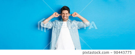 Irritated man close ears from noisy neighbours, looking bothered while standing against blue background Irritated man close ears from noisy neighbours, looking bothered while standing against blue background 98443775