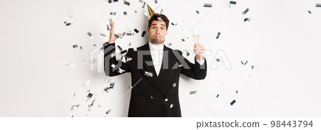 Handsome party guy in black suit having fun, celebrating new year, blowing whistle and drinking champagne while confetti falling, wishing happy holidays, standing against white background 98443794
