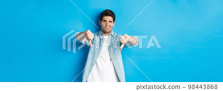 Disappointed man showing thumbs down and grimacing at something awful, dislike product, judging bad advertisement, standing against blue background Disappointed man showing thumbs down and grimacing at something awful, dislike product, judging bad advertisement, standing against blue background 98443868