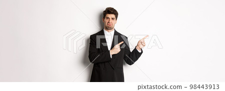 Portrait of disappointed and sad handsome businessman in suit, complaining and pointing fingers left at something bad, standing upset against white background Portrait of disappointed and sad handsome businessman in suit, complaining and pointing fingers left at something bad, standing upset against white background 98443913